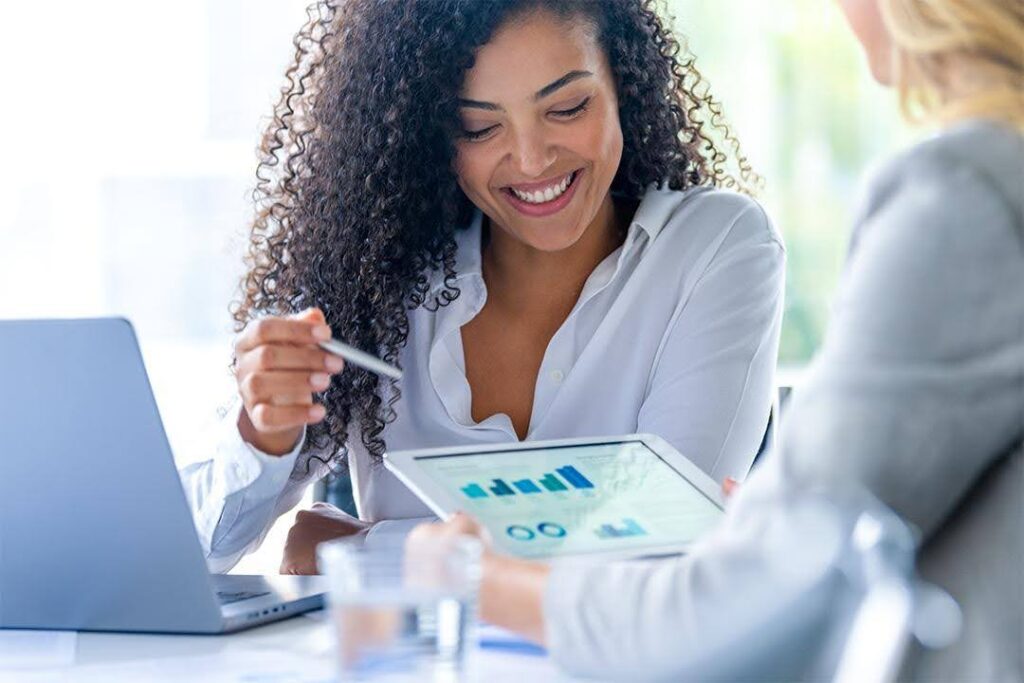Two businesswomen seated at a table discuss data on a tablet; one has a laptop open.