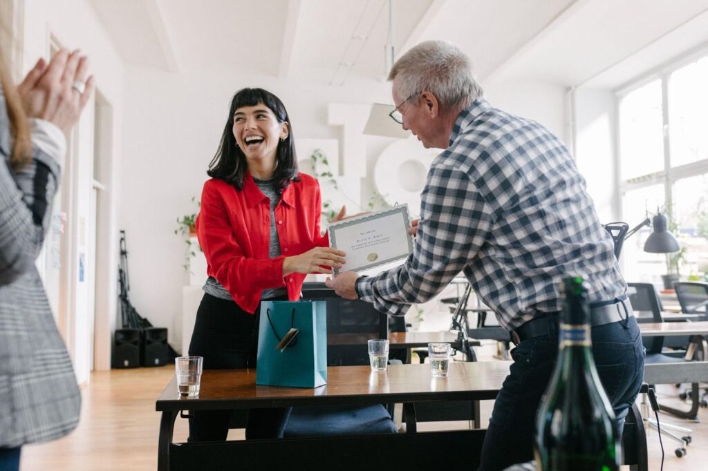 A smiling woman receives a certificate from an older man in an office setting, with a gift bag and drinks on the table and colleagues clapping nearby.