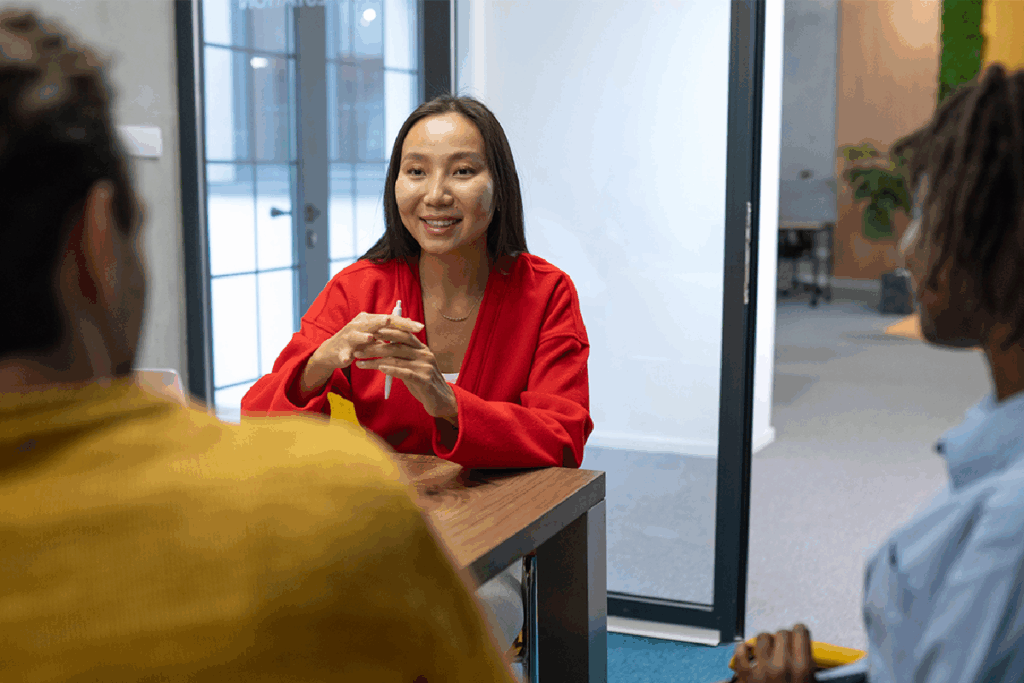 A smiling woman in a modern office setting with glass walls and an open workspace talks with two people across a table.