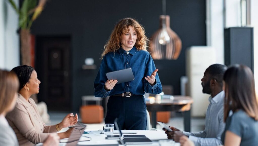 A woman holding a tablet stands and speaks to a group of colleagues seated around a table in a modern office meeting room.