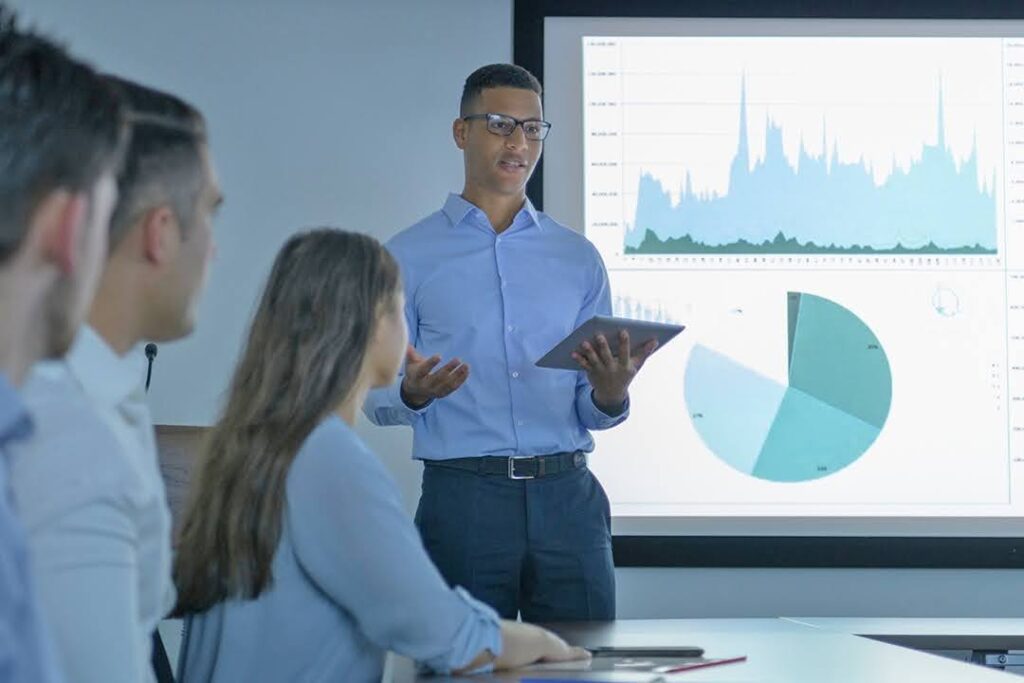 A man holding a tablet presents data with charts and graphs on a screen to a group of colleagues in a conference room.