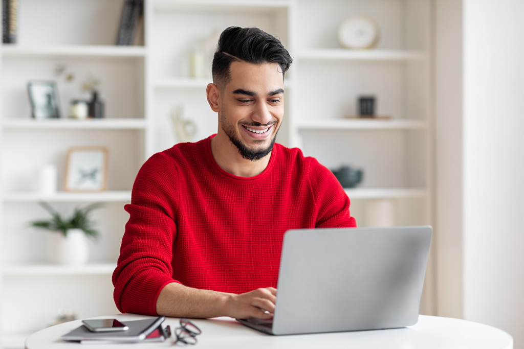 A smiling man in a red sweater uses a laptop at a table.