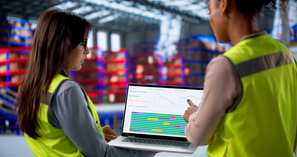 Two individuals in yellow vests analyze logistics data on a laptop in a warehouse with stacks of blue and red containers in the background.