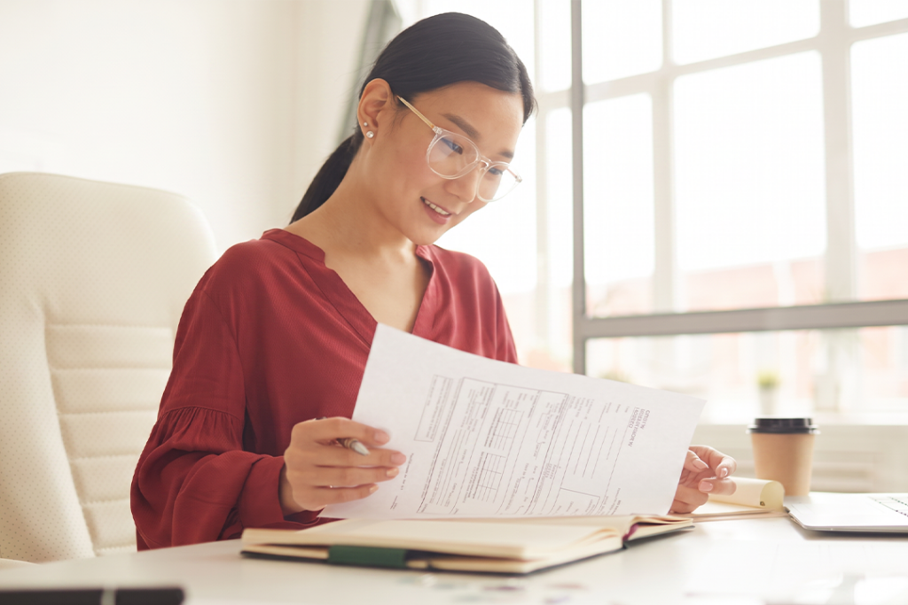 A woman wearing glasses reviews documents at a bright office desk.